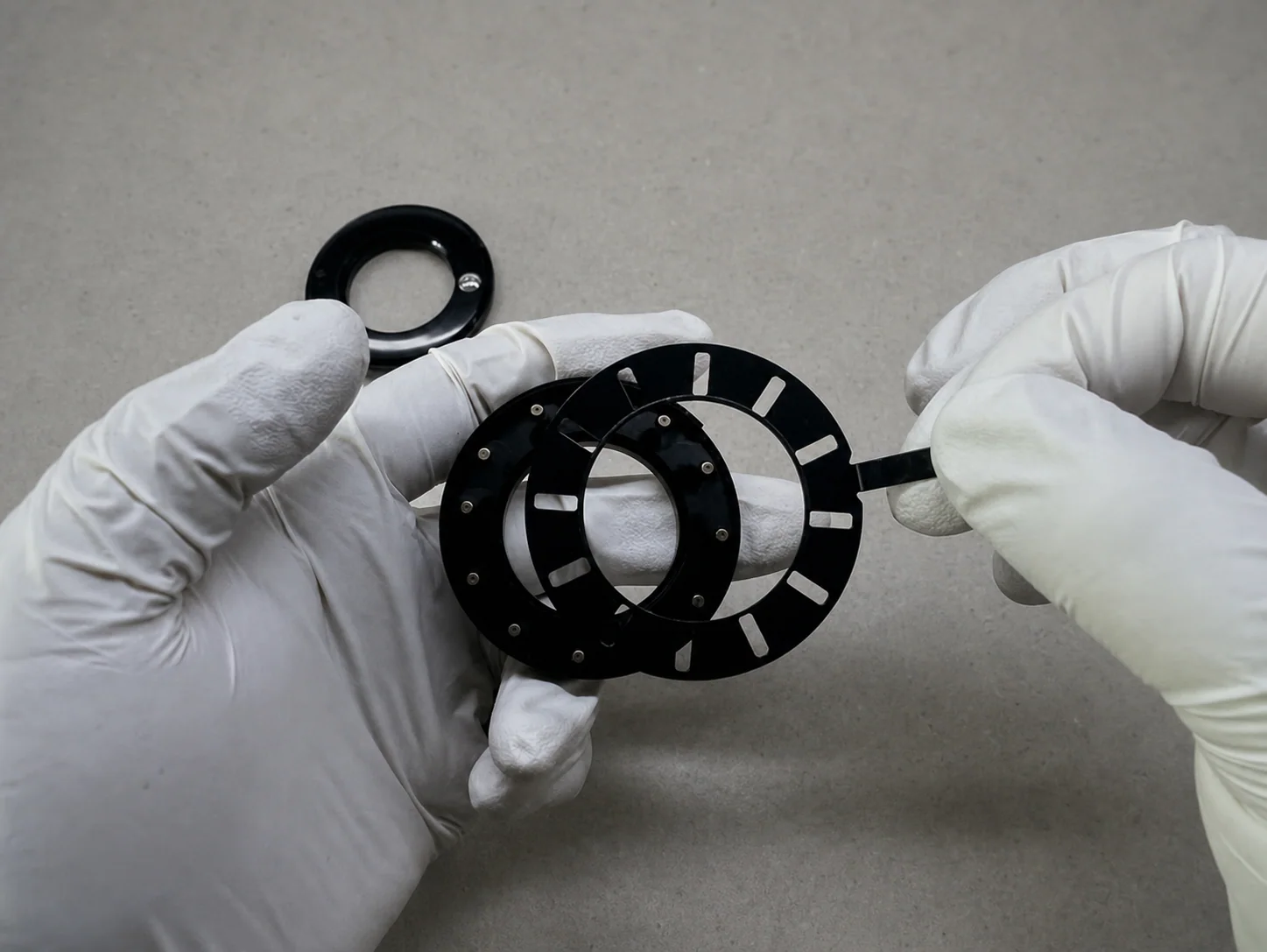 Gloved hands positioning black iris blade-mechanism components on a gray workbench